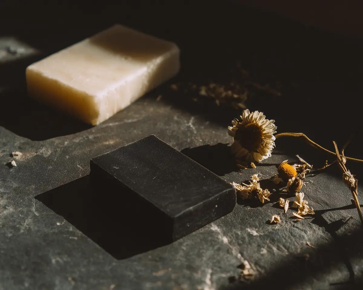 Schwarzes Syndet-Stück und heller Seifenriegel auf dunklem Stein, dazwischen getrocknete Kamillenblüten
