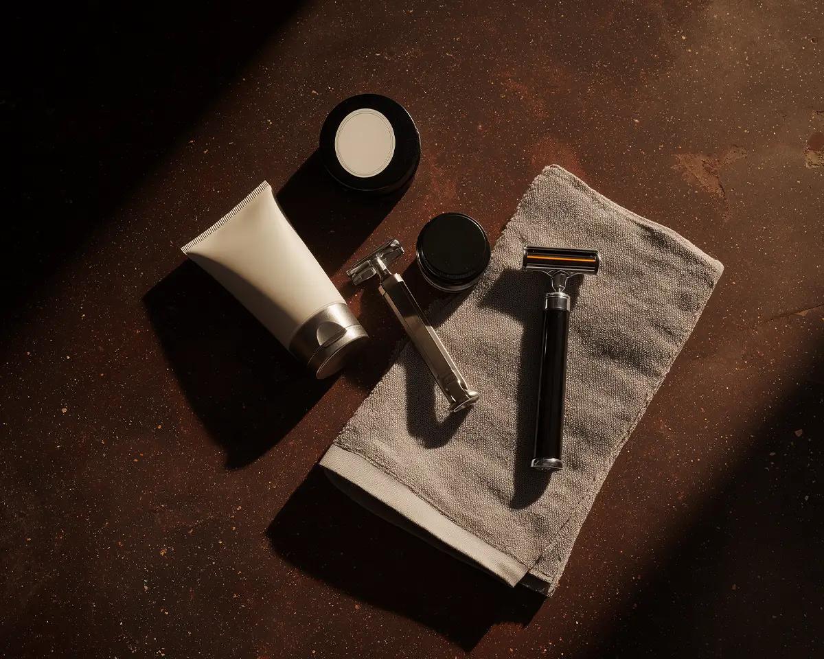 Draufsicht auf Rasierzubehör auf Leinentuch: Rasierhobel, weiße Creme-Tube, schwarzer Syndet-Puck und rundes Pflegeprodukt auf dunklem Untergrund